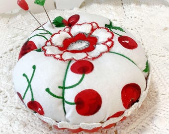 May include: A round fabric pincushion featuring a white background with a red cherry print and green stems. The top center is decorated with a white and red embroidered flower applique. Two sewing pins with red and green heads are inserted.