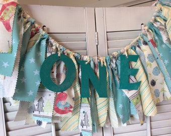 May include: A teal fabric banner with the word "ONE" in large teal letters. The banner is decorated with various patterns of fabric in shades of blue, green, yellow, and white.