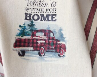 May include: A red and black plaid pickup truck with a Christmas tree in the back. The truck is driving through a snowy winter scene. The text "Winter is the time for home" is printed above the truck.
