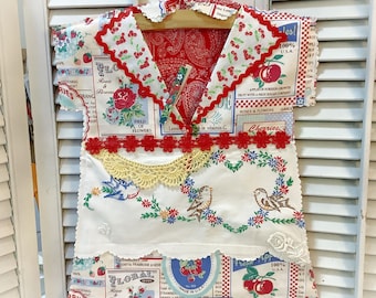 May include: A handmade dress crafted from vintage-style fabric featuring a white base with colorful floral, cherry, and bird embroidery. The garment includes a red cherry-patterned collar, a yellow lace accent, and red floral trim details.