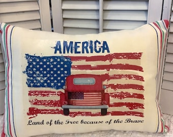 May include: A white pillow with a red, white, and blue American flag design. The flag is distressed and has a vintage look. A red truck with a white star on the back is in front of the flag. The text "AMERICA Land of the Free because of the Brave" is printed on the pillow.
