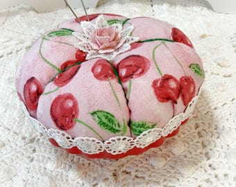 May include: A round pincushion featuring a light pink fabric with a red cherry and green leaf print. The cushion is accented with a white lace trim around the base, a white lace flower detail on top, and a small pink fabric rose in the center.
