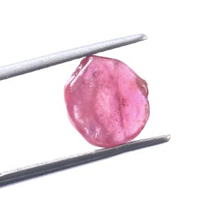 Puede incluir: Una gema rosa sujeta con pinzas plateadas sobre un fondo blanco. La gema tiene una forma lisa e irregular y una calidad translúcida, con variaciones de color.