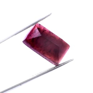 May include: A rectangular, faceted gemstone with a deep burgundy color is held by silver tweezers against a white background. The gem's edges are sharply defined, and its surface reflects light, showcasing its clarity and cut.