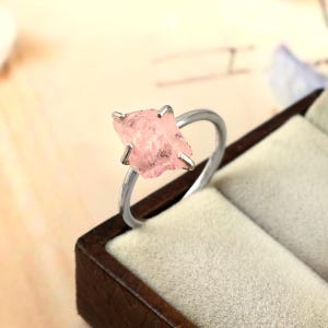以下が含まれることがあります： 4本の金属製の爪で固定された、未加工の淡いピンク色の宝石が特徴のシルバー調の指輪。石は不規則なファセット加工の表面テクスチャを持ち、滑らかで細い円形の金属バンドに取り付けられています。
