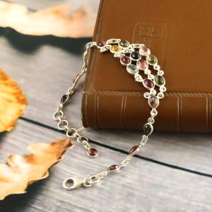 May include: A silver bracelet featuring a series of oval gemstones in shades of pink, green, yellow, and black. The bracelet is displayed on a brown leather surface and a wooden background.