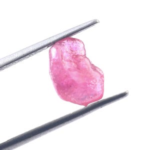 May include: A rough, pink gemstone is held by a pair of silver tweezers against a white background. The gemstone has an irregular shape and a slightly translucent appearance, with variations in color.
