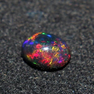 May include: A polished, oval-shaped black opal gemstone. The opal displays a vibrant play-of-color, with flashes of red, orange, yellow, green, and blue. The gemstone is set against a dark, textured background.
