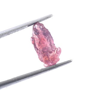 May include: A rough, uncut, pink gemstone is held by a pair of silver tweezers against a white background. The gemstone has a textured surface and varying shades of pink and red.
