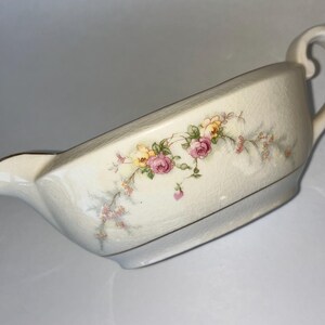 Pode incluir: Uma molheira de porcelana branca com um design floral com rosas cor-de-rosa, amarelas e brancas. A molheira tem uma alça do lado direito e um bico do lado esquerdo.