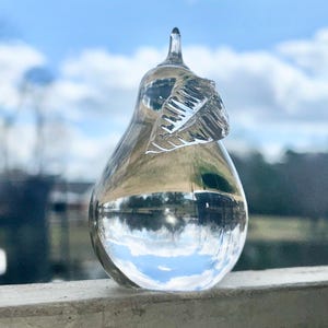 Pode incluir: Uma escultura de pêra de vidro transparente com um design de folha detalhado. A pêra reflete o céu e as nuvens.