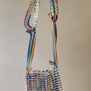 May include: A handmade, rainbow-coloured purse made from recycled aluminium can tabs. The bag features a rectangular body and a strap made of can tabs and rainbow ribbon. The bag is suspended from a metal hook.