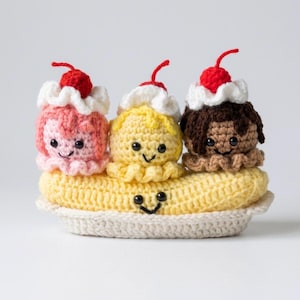 May include: A crocheted banana split toy featuring three scoops of ice cream in pink, yellow, and brown, each topped with white whipped cream and a red cherry. The scoops and the yellow banana base have embroidered black eyes and smiling mouths.