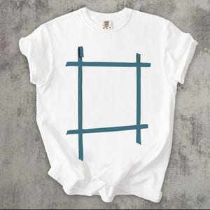 May include: A white short-sleeved t-shirt featuring a central graphic of a dark teal square frame design. The graphic consists of four intersecting lines that form a simple, open-ended rectangular shape. The shirt has a crew neckline and a label.