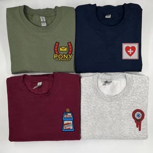 Puede incluir: Cuatro sudaderas de diferentes colores con diseños bordados. La primera sudadera es de color verde oliva con una herradura amarilla y el texto "PONY EXPRESS". La segunda sudadera es azul marino con una cruz roja en un cuadrado blanco. La tercera sudadera es de color burdeos con una botella de solución de limpieza de dibujos animados azul y blanca. La cuarta sudadera es de color gris claro con un ojo rojo con una pupila blanca y una gota roja.