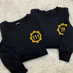 May include: Two black sweatshirts featuring yellow gear logos. One displays the number '111' within the gear, the other '76'. The garments are folded, presented on a white, textured surface.