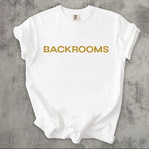 May include: A white short-sleeved t-shirt featuring the word BACKROOMS printed in a bold, gold-coloured sans-serif font across the centre of the chest. The garment has a crew neckline and a small brand label visible at the inside of the collar.