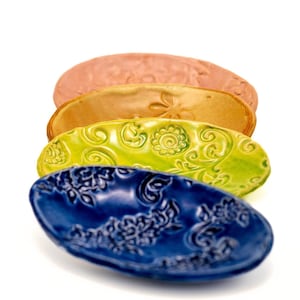 Puede incluir: Cuatro fuentes de cerámica en fila, cada una con un color y un diseño en relieve únicos. Los colores son, de arriba a abajo, rosa empolvado, bronce dorado, verde brillante y azul profundo. Los diseños incluyen patrones florales y abstractos.