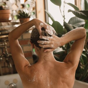K&ouml;nnte beinhalten: Eine Person mit nassem Haar w&auml;scht ihre Haare unter der Dusche. Sie verwendet ihre H&auml;nde, um Shampoo oder Conditioner aufzusch&auml;umen. Die Person steht in einem Badezimmer mit einer gekachelten Wand und einer Pflanze im Hintergrund.