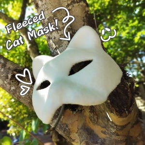 Può includere: Una maschera da gatto in pile bianco con fori per gli occhi e forma di orecchie. La maschera è esposta su un ramo d'albero. Il testo sull'immagine recita "Fleeced Cat Mask!"