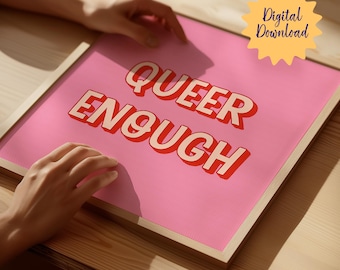 May include: A pink framed print with the text "QUEER ENOUGH" in red and white letters. The print is on a wooden table.