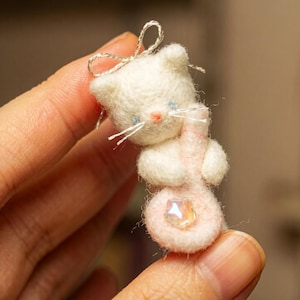 Könnte beinhalten: Eine kleine gefilzte Katzenfigur in Weiß mit einer silbernen Schleife auf dem Kopf, die ein hellrosa gefilztes Objekt mit einem klaren, irisierenden Kristallakzent hält. Die Katze hat blaue Augen, eine rosa Nase und weiße Schnurrhaare.
