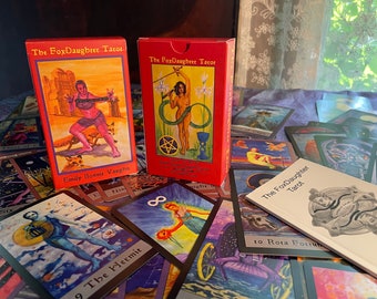 May include: A collection of tarot cards and boxes. The boxes are red with the text "The FoxDaughter Tarot" and illustrations. Several tarot cards are spread out, featuring various artistic depictions and titles like "The Hermit" and "Rota Fortuna".