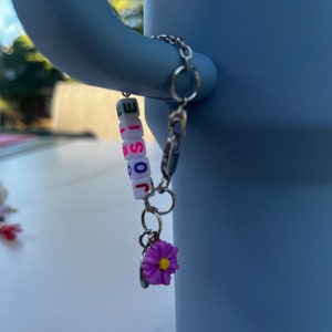 May include: A silver chain with a charm that spells out "JOSIE" in colorful letter beads. The charm also has a small purple flower attached to it.