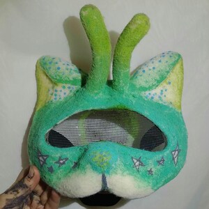 May include: A handmade green and white mask with a mesh eye covering. The mask features two green antennae, cat-like ears, and star and floral designs. The mask is made of a soft, textured material.