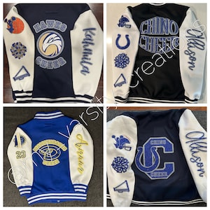 May include: Four varsity cheerleading jackets in navy, black, and blue with white sleeves. Each jacket features team names, cheerleading graphics, and personalized names in glittery lettering. The jackets have striped cuffs and collars.