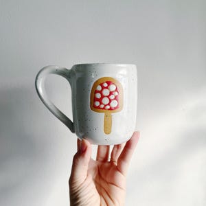 May include: White ceramic mug with a speckled finish, featuring a whimsical mushroom design. The mushroom has a red cap with white spots and a tan stem. The mug is held by a hand.