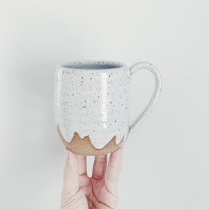 May include: Handmade ceramic mug with a speckled white glaze and a brown base. The mug has a curved handle and a unique design where the white glaze drips down the sides. A perfect addition to any kitchen or coffee collection.