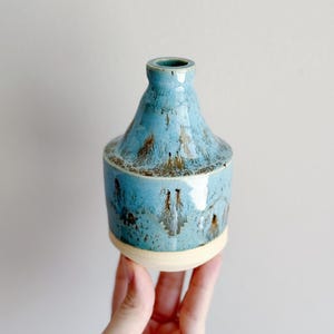 May include: A handmade ceramic vase with a unique shape and a striking blue glaze. The vase features a wide base, a rounded body, and a narrow neck. The blue glaze has brown and gold accents, adding depth and visual interest. The vase is held in a hand.