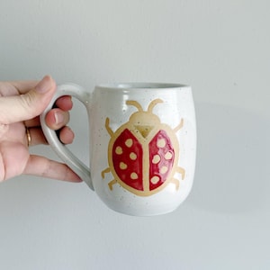 May include: A white ceramic mug with a speckled finish featuring a hand-painted ladybug design. The ladybug has a tan body with red wings and dark red spots. The mug has a curved handle and a rounded body shape, providing a sturdy grip for use.