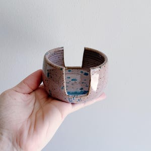 May include: A handmade ceramic napkin holder in a speckled mauve color with teal blue accents. The holder has a circular shape with two cut-out sections. The interior is glazed, and the exterior has a textured finish.