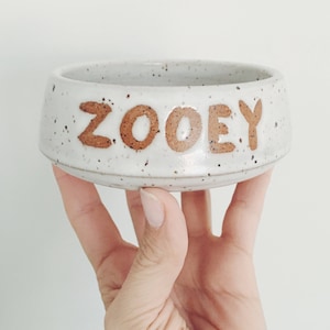 May include: A speckled white ceramic pet bowl with the name "ZOOEY" in brown lettering. The bowl is held by a hand, showcasing its round shape and low profile. The bowl is designed for pets.