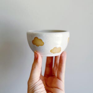 May include: A small, off-white ceramic bowl with a rounded shape. The bowl is decorated with several light brown cloud shapes. The bowl is held in a hand against a plain, light gray background. The bowl has a speckled texture.