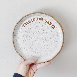 May include: A white ceramic plate with a speckled design and a brown rim. The words "TREATS FOR SANTA" are written in brown lettering across the top of the plate. The plate is held by a hand.