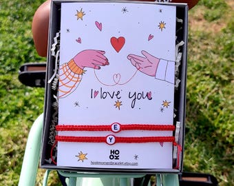 Puede incluir: Pulseras de cuerda roja con abalorios de letras blancas que deletrean "YOU" y "YE" en una caja de regalo con una tarjeta que dice "Te amo" con una pareja de dibujos animados tomándose de la mano y un corazón rojo.