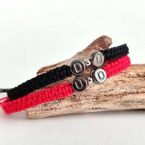 May include: Two macrame bracelets, one black and one red, are displayed on a piece of driftwood. Each bracelet features a silver charm with the letters "D" and "U". The bracelets are knotted with a sliding knot closure.
