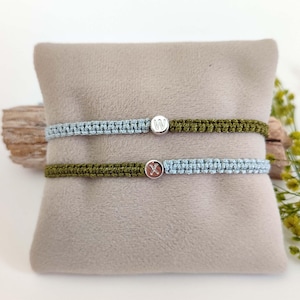 May include: Two friendship bracelets with silver charms. The top bracelet is green and blue, with a silver charm that says "W". The bottom bracelet is green and blue, with a silver charm that says "X".