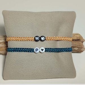以下が含まれることがあります： 黒と白のビーズが付いた友情ブレスレット2本。上のブレスレットは金色で、「W」と「♥」の文字が書かれています。下のブレスレットはターコイズブルーで、「P」と「♥」の文字が書かれています。