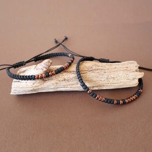 Op de afbeelding: Twee zwarte gevlochten koordarmbanden met koperkleurige kralen. De armbanden zijn verstelbaar met schuifknopen en worden gepresenteerd op een stuk drijfhout tegen een bruine achtergrond. De kralen zijn gelijkmatig verdeeld.