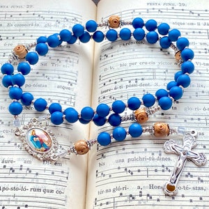May include: A blue rosary with wooden rose beads and a silver crucifix. The rosary features a detailed centerpiece with an image and is set against a backdrop of an open book with musical notes.