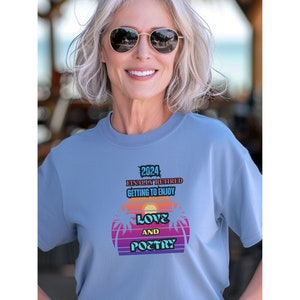 May include: A light blue t-shirt with a graphic design featuring a sunset with palm trees and the text "2024 Finally Retired Getting to Enjoy Love and Poetry".