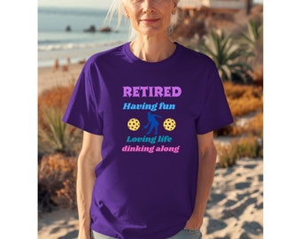 May include: Purple t-shirt with the text "RETIRED Having fun Loving life dinking along" and a silhouette of a person playing pickleball.