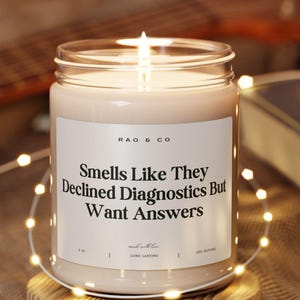 May include: A lit candle in a clear glass jar with a white label that reads "Smells Like They Declined Diagnostics But Want Answers." The candle sits on a small wooden coaster, surrounded by string lights. A guitar and a book are in the background.