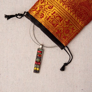 Puede incluir: Un collar de plata con un colgante de mosaico de colores cuelga de una bolsa de tela roja y dorada con estampado.