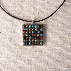 May include: A square pendant necklace with a black leather cord. The pendant is made of silver and features a mosaic of colorful gemstones.