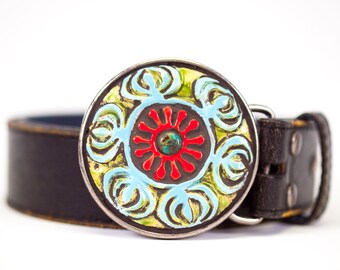 May include: A black leather belt with a large, round, silver buckle. The buckle is decorated with a colourful, geometric design featuring blue, green, red, and yellow.